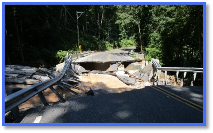 "A paved roadway in a wooded area is severely damaged, with a large section collapsed and washed out. Guardrails on both sides are bent and broken near the damaged area. Trees and vegetation surround the roadway, and utility poles line the left side of the road."